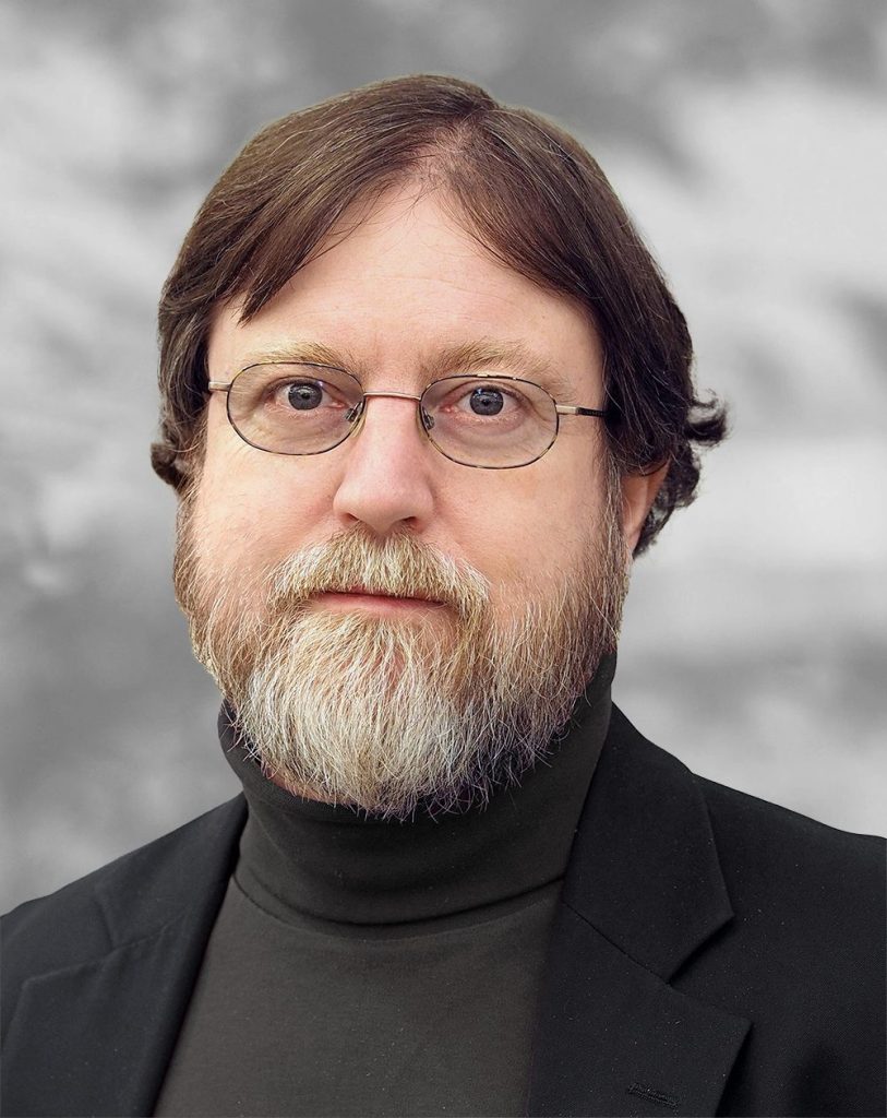 A portrait of Augustus Lowell, a man with glasses and a graying beard wearing a bark turtleneck against a mottled gray background.