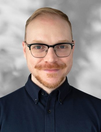 A portrait of Daniel Badali, a smiling man with glasses and a beard against a gray background.