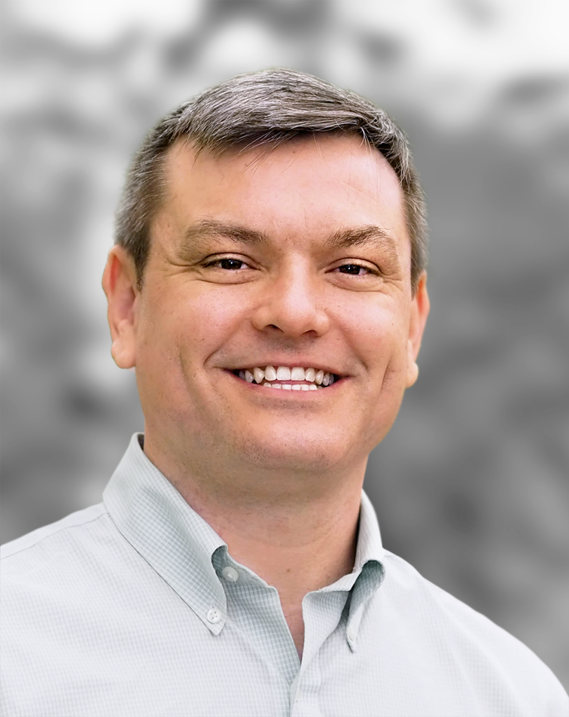 A portrait of Jeffrey Perkins, a smiling man in a buttoned shirt with a gray background.