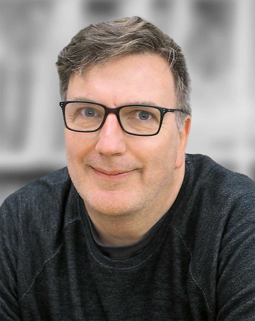 A portrait of Joe Heanue, a smiling man with glasses against a gray background.