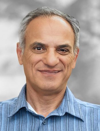 A portrait of Mansour Saleki, a smiling man with graying hair wearing a blue striped shirt against a blurred gray background.