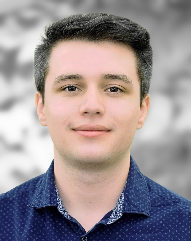 A portrait of Nicholas Mann, a man with a neutral expression, wearing a blue patterned shirt against a gray background.