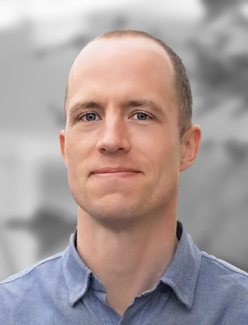 A portrait of Robert de Saint Phalle, a smiling man wearing a blue shirt, with a blurred background.