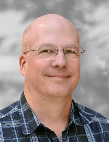 A portrait of Roger Stenerson, a smiling man wearing glasses and a plaid shirt against a blurred background.