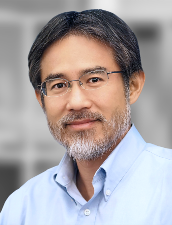 A portrait of Roger Tang, a smiling man with gray hair, a beard, and glasses, wearing a blue shirt, against a blurred background.