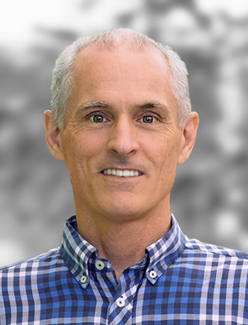 A portrait of Ryan McGuinness, a smiling man wearing a checkered shirt, against a blurred background.