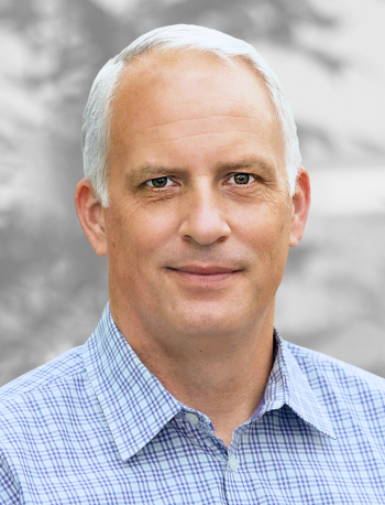 A portrait of Tim Buskard, a smiling man with short white hair, wearing a blue checkered shirt, against a blurred background.