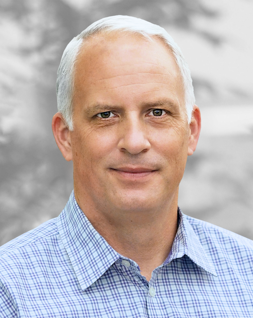 A portrait of Tim Buskard, a smiling man with short white hair, wearing a blue checkered shirt, against a blurred background.