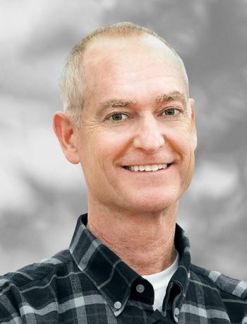 A portrait of of Walt Cecka, a smiling man with short hair wearing a plaid shirt against a blurred background.