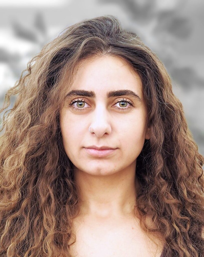 A portrait of Lilit Karayan, a woman with long curly hair and green eyes is looking directly at the camera with a neutral expression. The background is blurred.