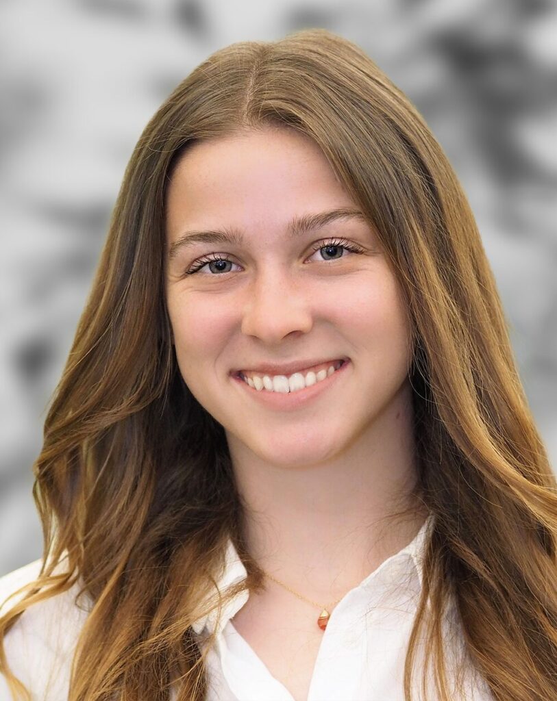 Portrait of Cameran Casale, a person with long, straight brown hair, wearing a white shirt and a necklace, smiling against a blurred background.