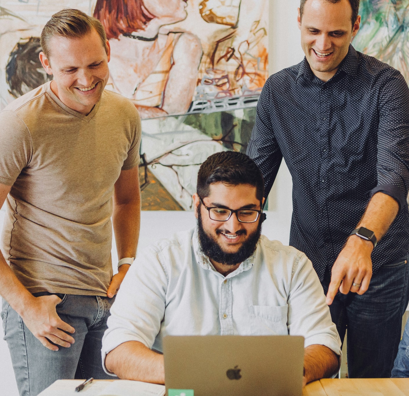 Three men smiling while looking at a laptop screen in a casual office setting.